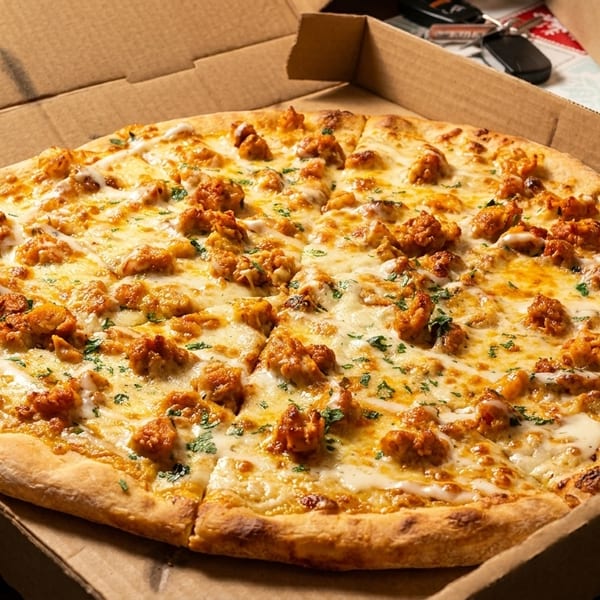 A whole pizza with white sauce, melted cheese, and chicken pieces in a cardboard box.
