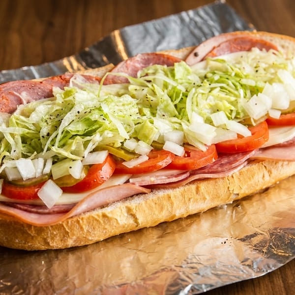 A large Italian sub sandwich with various meats, cheese, lettuce, tomatoes, onions, and pickles on foil.