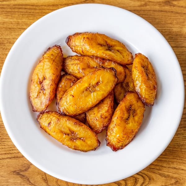 A white plate filled with golden-brown fried plantains, glistening on a wooden table.